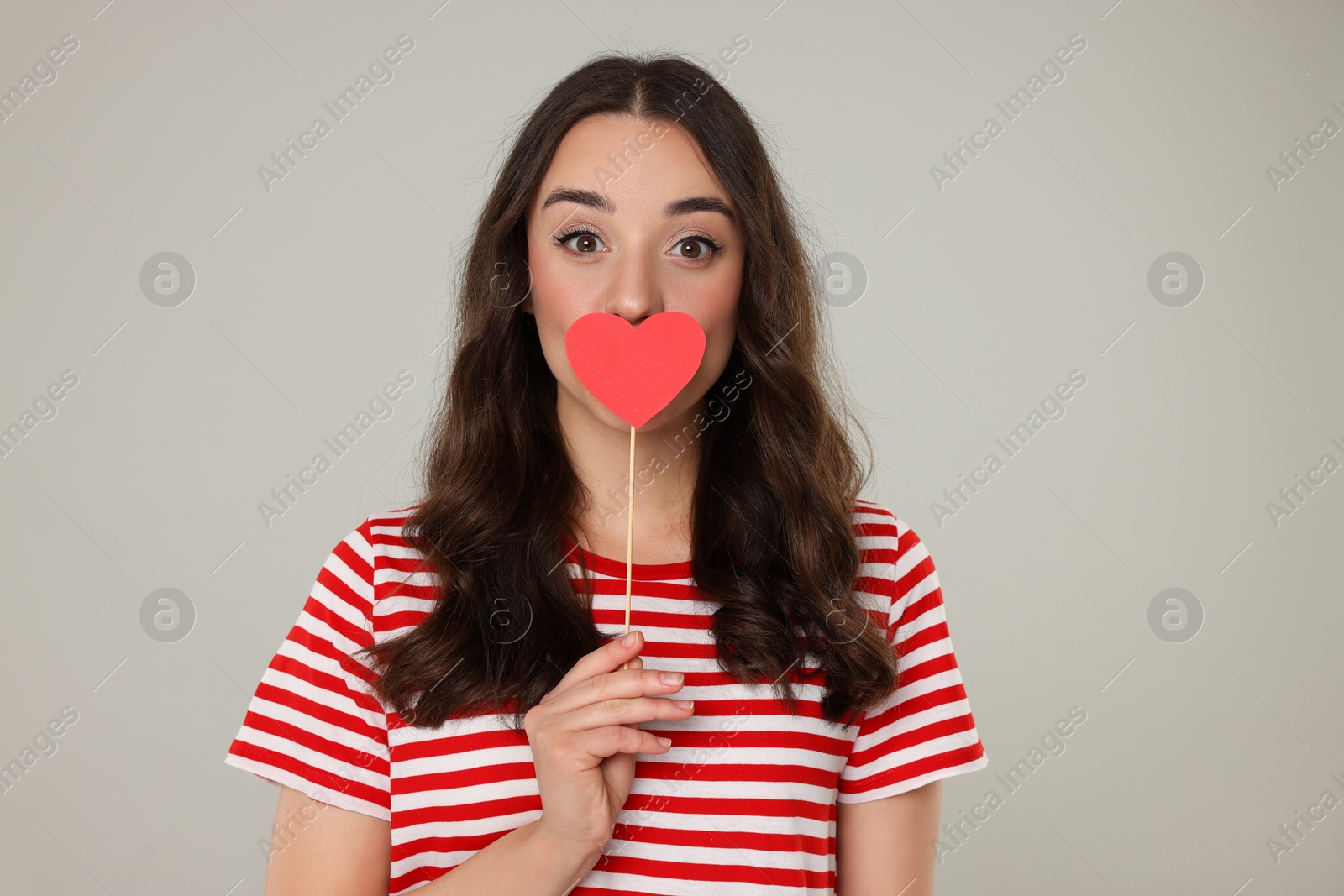 Photo of Beautiful young woman covering her mouth with paper heart on grey background