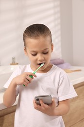 Photo of Little girl using smartphone while brushing teeth in bathroom. Internet addiction