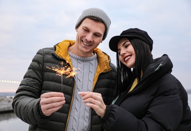Photo of Couple in warm clothes holding burning sparklers near river