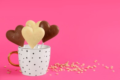 Photo of Heart shaped lollipops made of chocolate in cup and sprinkles on pink background, space for text