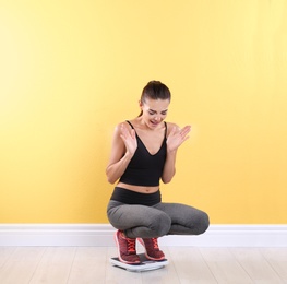 Emotional young woman measuring her weight using scales near color wall. Weight loss motivation