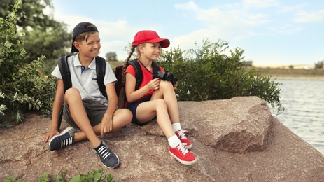 Image of Cute little children with backpacks sitting on rock near river. Camping trip