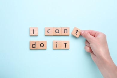 Motivation concept. Woman changing phrase from I Can't Do It into I Can Do It by removing wooden square with letter T on light blue background, top view