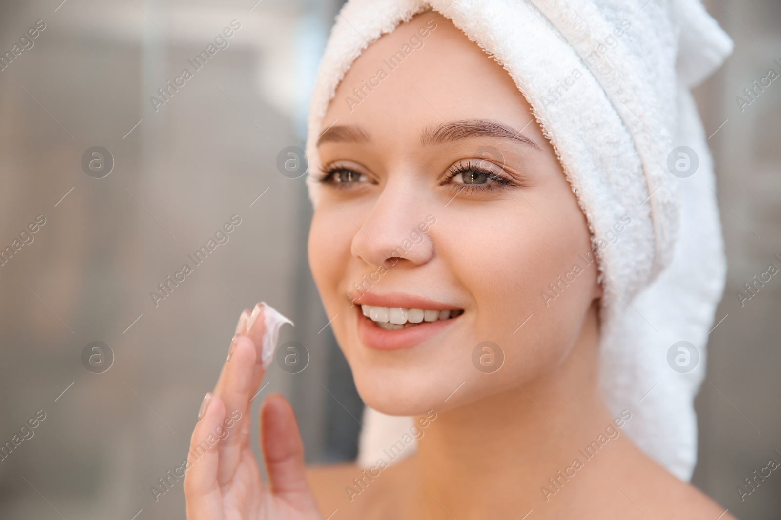 Photo of Beautiful woman with towel on head applying face cream in bathroom