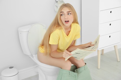 Young woman reading newspaper while sitting on toilet bowl at home