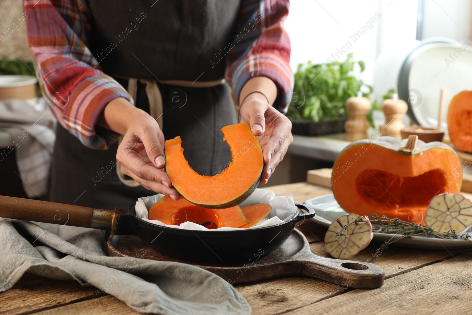 Photo of Woman putting pumpkin slices into frying pan at wooden table in kitchen, closeup