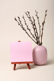 Photo of Vase with beautiful blooming willow branches and blank card on stand against beige background. Space for text