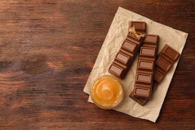 Tasty chocolate bars and bowl of caramel on wooden table, top view. Space for text