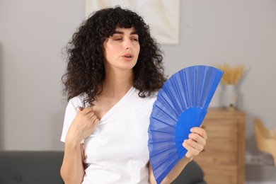 Young woman waving blue hand fan to cool herself at home