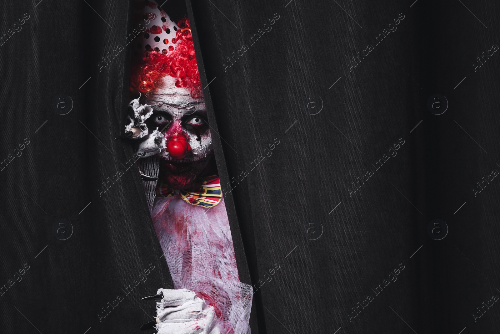 Photo of Terrifying clown hiding behind black curtains, space for text. Halloween party costume