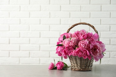 Photo of Wicker basket with fragrant peonies on table against brick wall, space for text. Beautiful spring flowers