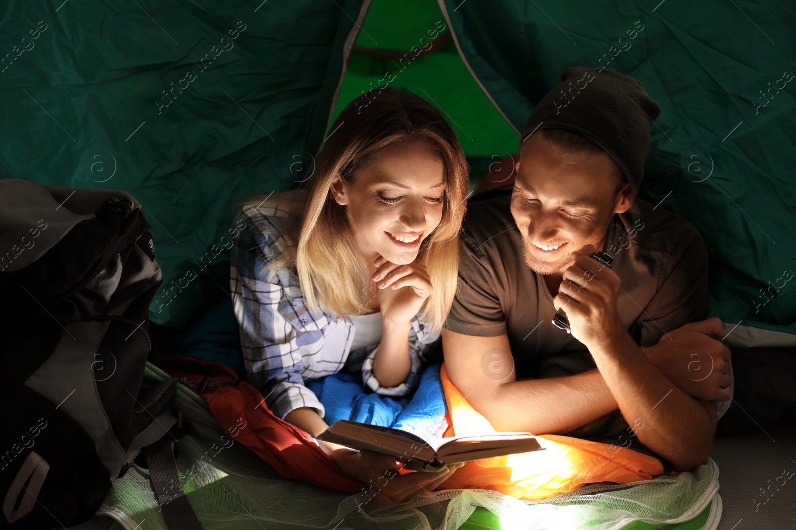 Photo of Young couple in sleeping bags reading book with flashlight inside of tent