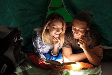 Photo of Young couple in sleeping bags reading book with flashlight inside of tent