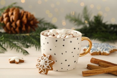 Photo of Delicious hot chocolate with marshmallows, gingerbread cookies and cinnamon on white wooden table against blurred festive lights