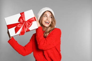 Happy young woman in Santa hat and sweater with gift box on light grey background. Christmas celebration