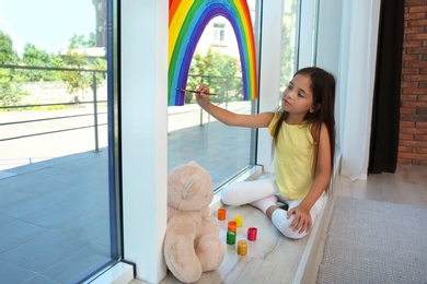 Photo of Little girl drawing rainbow on window with paints indoors. Stay at home concept