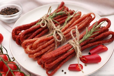 Photo of Bundles of delicious kabanosy with rosemary, peppercorn, chilli and tomatoes on white wooden table, closeup