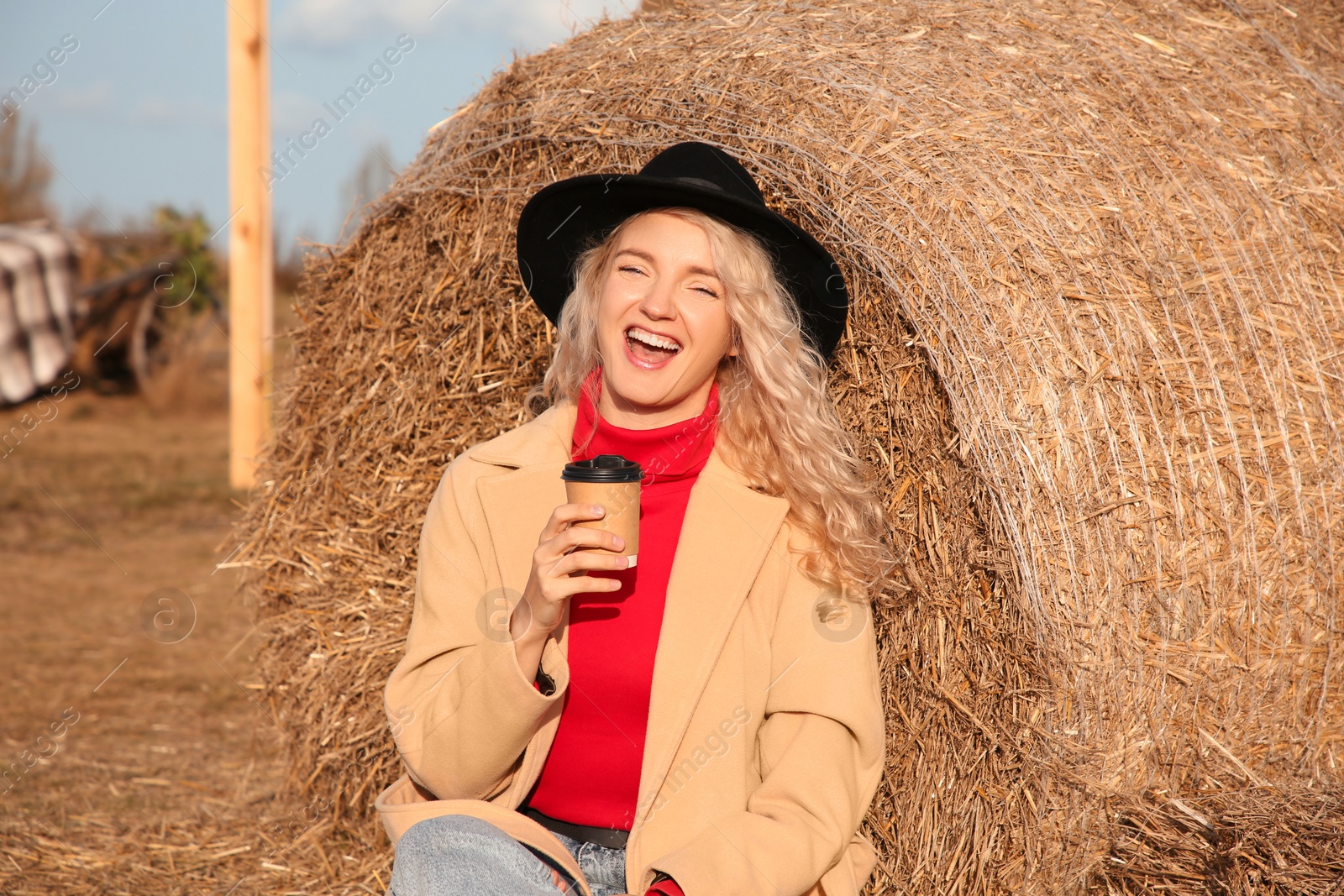 Photo of Happy woman with cup of hot drink sitting near hay bale outdoors. Autumn season