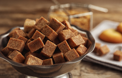 Photo of Brown sugar cubes in metal bowl on wooden table, closeup