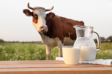 Photo of Glass with jug of milk on wooden table and cow grazing in meadow