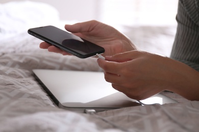 Photo of Woman connecting charger cable to smartphone near laptop on bed, closeup