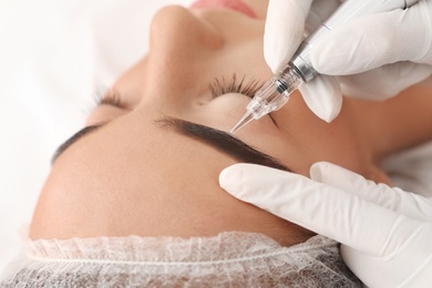 Young woman undergoing procedure of permanent eyebrow makeup in tattoo salon, closeup