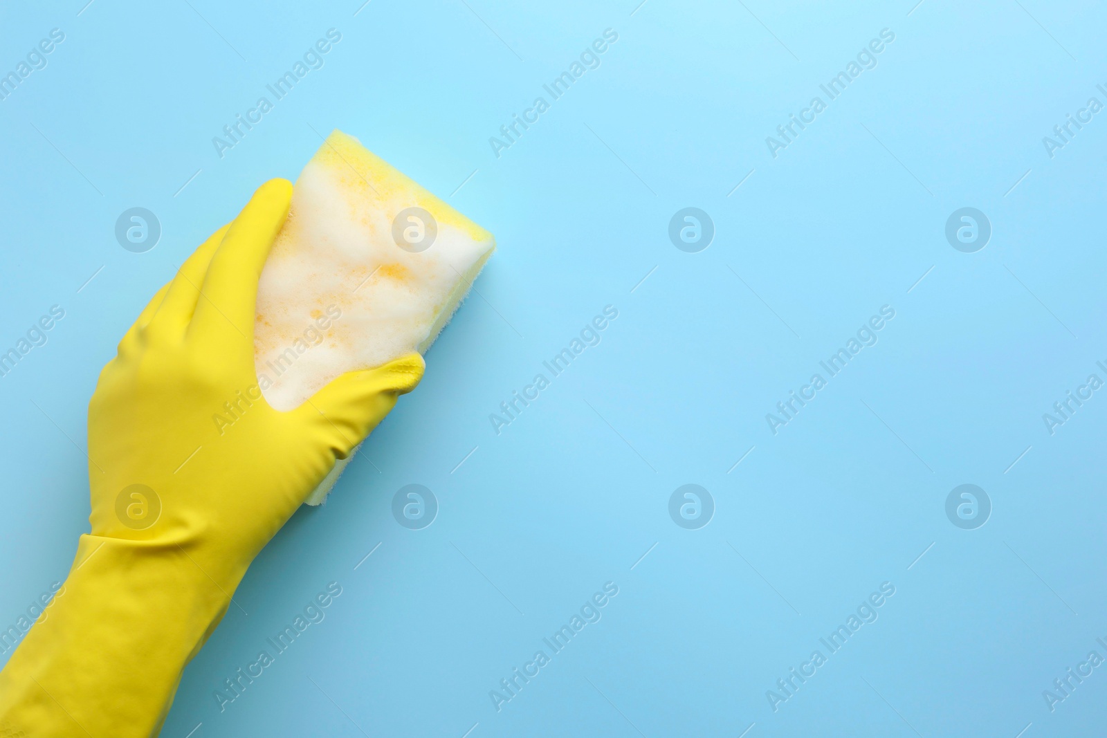 Photo of Cleaner in rubber glove holding sponge with foam on light blue background, top view. Space for text