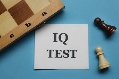 Photo of Note with text IQ Test, chess board and pieces on light blue background, flat lay
