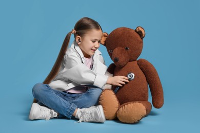 Little girl in medical uniform examining toy bear with stethoscope on light blue background