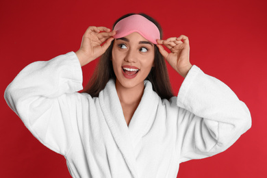 Photo of Young woman in bathrobe and eye sleeping mask on red background