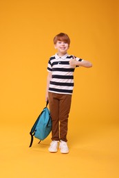 Photo of Smiling schoolboy with backpack showing thumb up on orange background