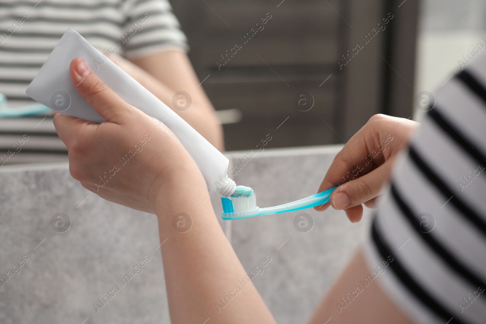 Photo of Woman squeezing toothpaste from tube onto toothbrush near mirror in bathroom, closeup