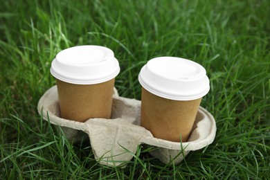 Photo of Takeaway paper coffee cups with plastic lids in cardboard holder on green grass outdoors