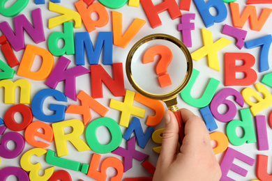 Woman holding magnifying glass over question mark surrounded by magnet letters on white background, top view. Search concept