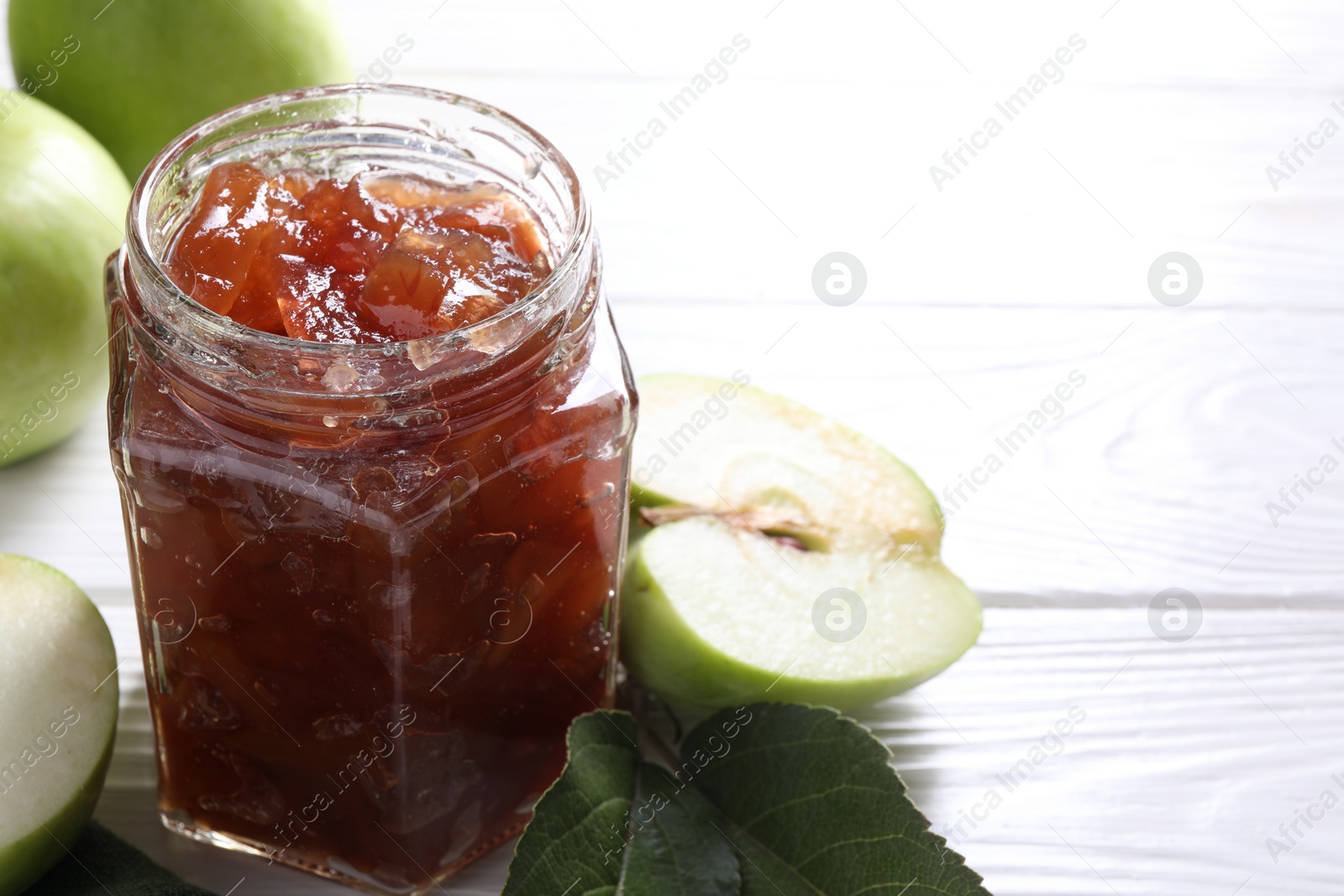 Photo of Glass jar of delicious apple jam and fresh fruits on white wooden table, closeup. Space for text