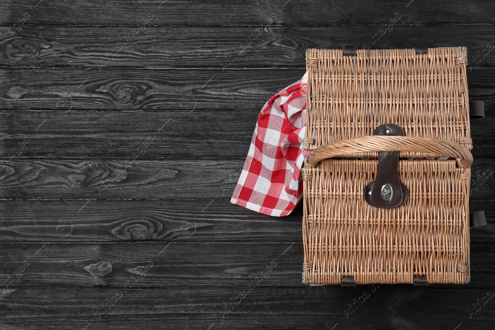 Photo of Checkered tablecloth and closed wicker picnic basket on black wooden table, top view. Space for text