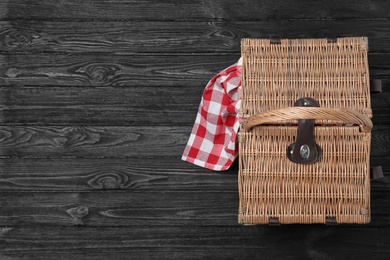 Photo of Checkered tablecloth and closed wicker picnic basket on black wooden table, top view. Space for text