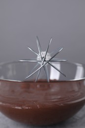 Whisk and bowl with chocolate cream on table against grey background, closeup
