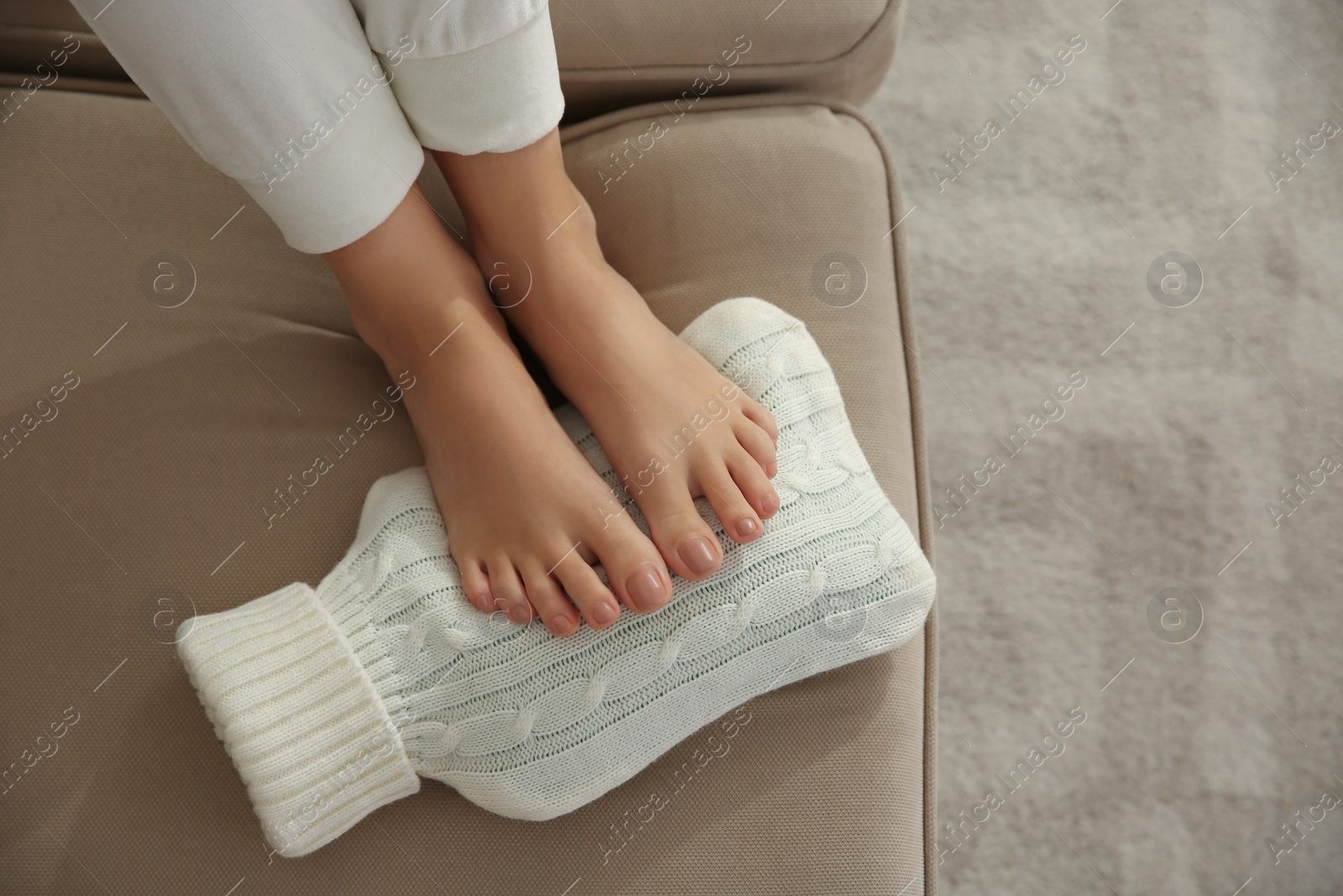 Photo of Woman warming feet with hot water bottle on sofa, closeup