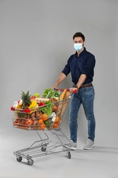 Young man in medical mask with shopping cart full of groceries on light grey background