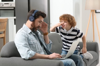 Little boy bothering his father at home. Man with laptop and headphones working remotely