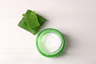 Jar of cosmetic cream and cut aloe vera leaves on white wooden table, top view