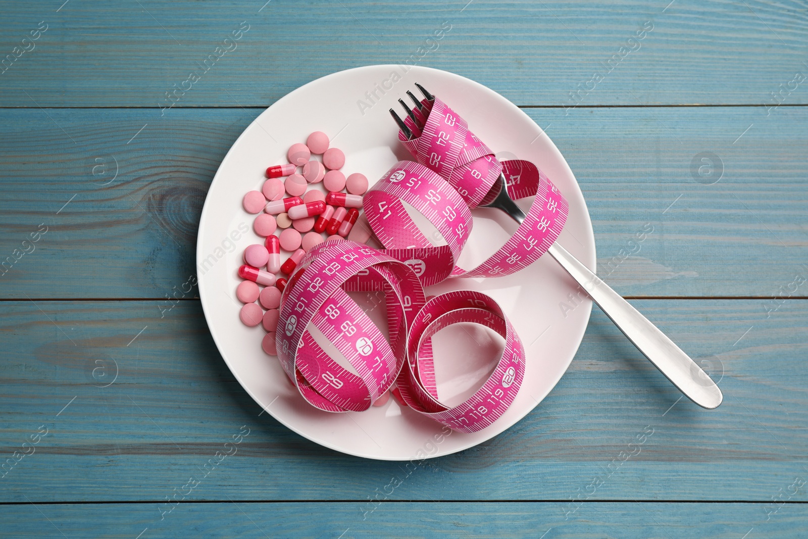 Photo of Plate with weight loss pills, measuring tape and fork on light blue wooden table, top view