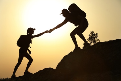 Silhouettes of man and woman helping each other to climb on hill against sunset