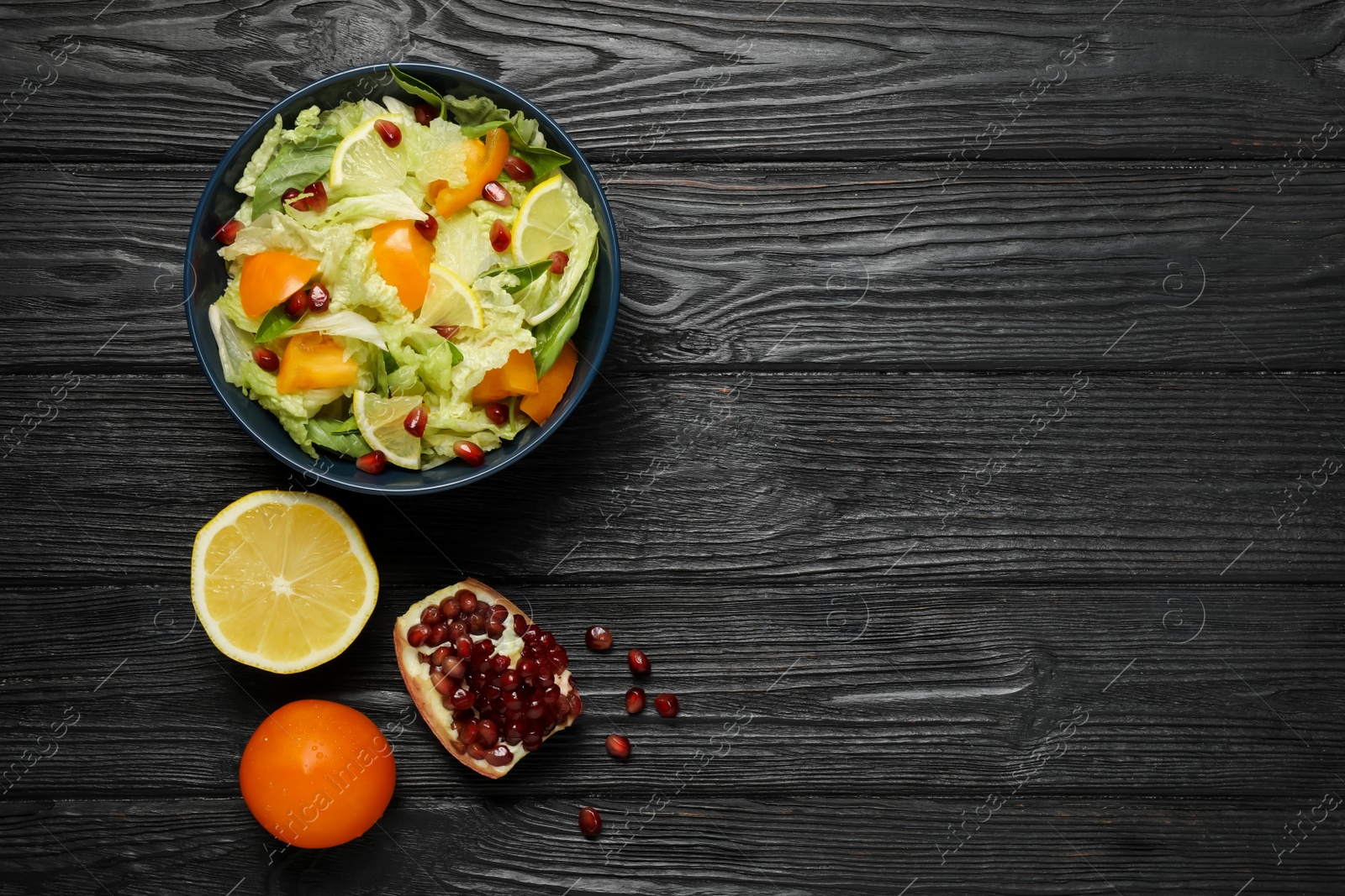 Photo of Delicious salad with Chinese cabbage, lemon, persimmon and pomegranate seeds on black wooden table, flat lay. Space for text