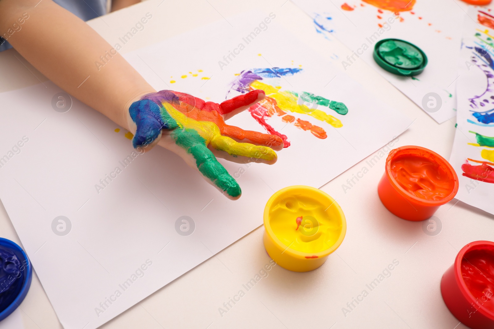 Photo of Little child making hand print on paper with painted palm at white table indoors, closeup