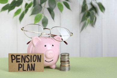 Photo of Card with phrase Pension Plan, piggy bank and stack of coins on light green table. Retirement concept