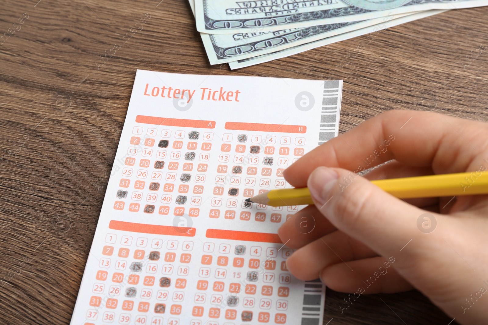 Photo of Woman filling out lottery ticket with pencil and money on wooden table, closeup
