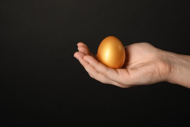 Photo of Woman holding shiny golden egg on black background, closeup. Space for text