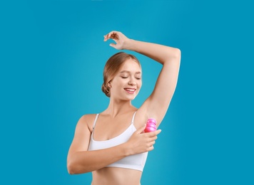 Young woman applying deodorant to armpit on teal background